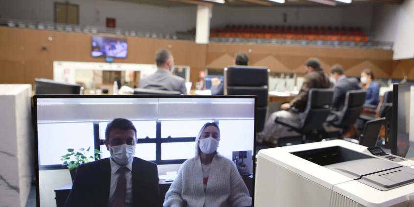 Monitor de computador exibe convidados em videoconferência. Ao fundo, vereadores compõem mesa de reunião no Plenário Amynthas de Barros. Galerias vazias mais ao fundo