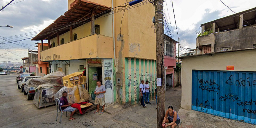 Beco à direita em rua com alguns carros cobertos ao fundo e casa com muro pixado à direita, durante o dia