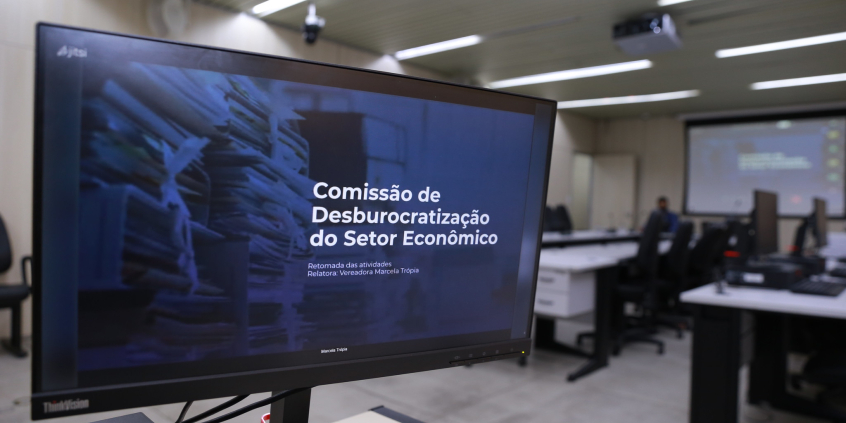 tela de computador em primeiro plano mostra nome da Comissão; a fundo, estrutura do Plenário Camil Caram