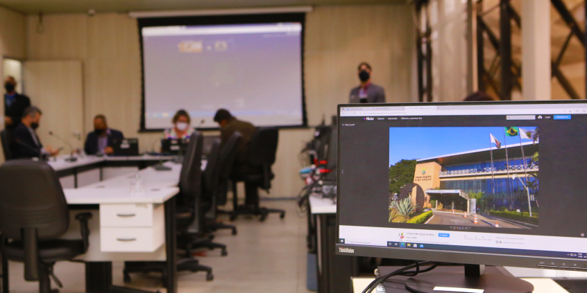 Reunião de Plenário realizada no plenário Helvécio Arantes. Monitor de computador exibe foto da fachada da Câmara. Ao fundo, vereadores compõem mesa de reunião. Telão exibe demais parlamentares em videoconferência