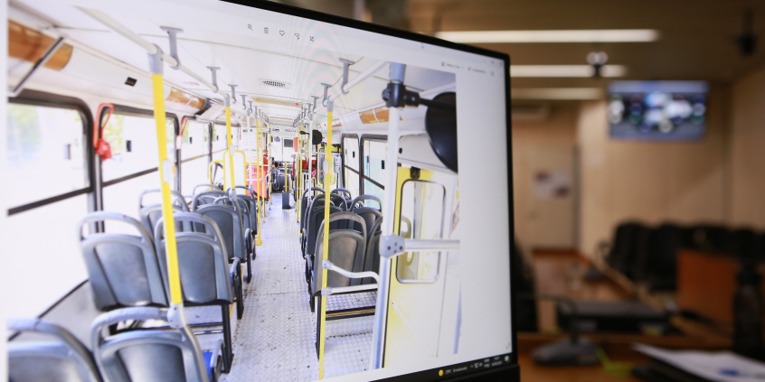tela de computador, em primeiro plano, mostra interior de um ônibus; ao fundo, mobília do Plenário Camil Caram 