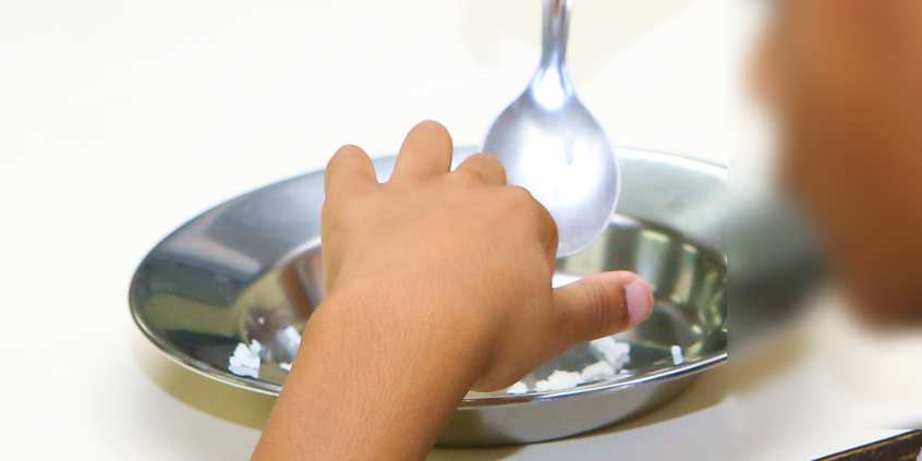 Criança se alimentando. À sua frente um prato de alumínio com pouca comida. A criança segura uma colher e usa a mão para manusear o alimento