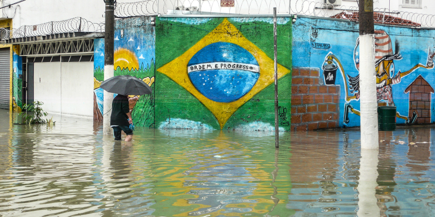 Imagem de uma pessoa atravessando com um  guarda-chuva, uma área totalmente alagada. Ao fundo, um muro com a bandeira do Brasil pintada. 