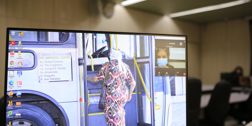 Imagem na tela do computador mostra idosa entrando em um ônibus. Ela tem os cabelos brancos e usa um vestido estampado e uma bolsa a tiracolo