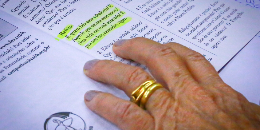 Mão feminina sobre publicação em que um refrão está sublinhado, com os seguintes dizeres E quem fala com sabedoria / É aquele que ensina com amor / Sua vida em total harmonia / É para nós luz, caminho, vigor”