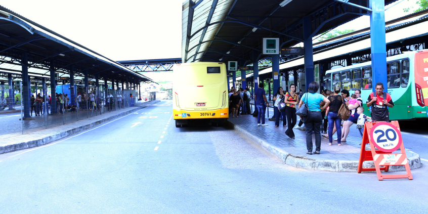 Na Estação Diamante passageiros, em pé, aguardam o ônibus. Estacionado, um ônibus amarelo aguarda a saída