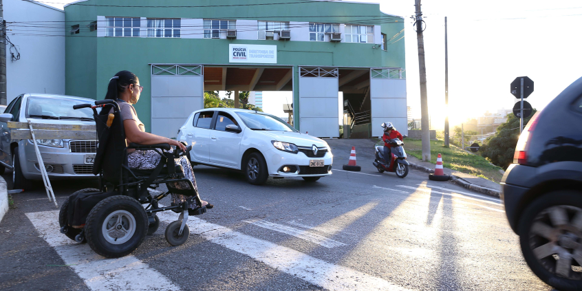 Cadeirante atravessa a rua em frente à Polícia Militar na faixa de pedestres, durante o dia. À esquerda, carro parado espera a travessia. 