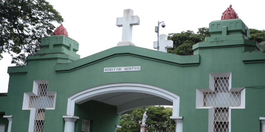 Entrada em arco do Cemitério do Bonfim. A entrada é pintada de verde e tem duas cruzes vazadas na edificação com detalhes em branco. Ao fundo, é possível avistar os túmulos
