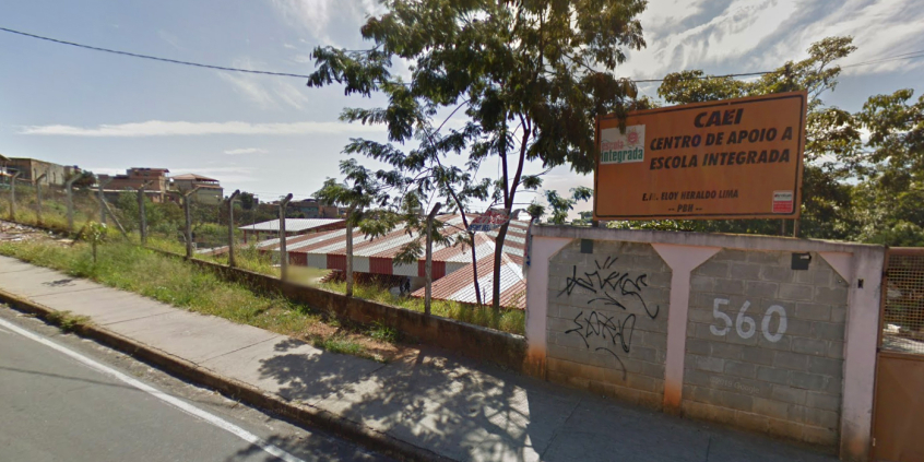 Terreno com grade e muro com placa com os seguintes dizeres: Caei - Centro de Apoio a Escola Integrada. Ao fundo, tenhado de residência, durante o dia. 
