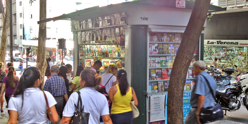 Várias pessoas passam diante de uma banca de revista no centro da cidade