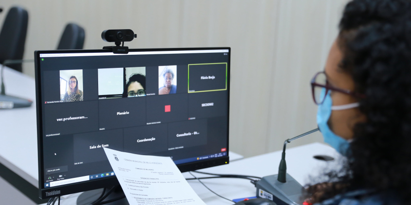 Vereadora, sentada à mesa, acompanha reunião remota em computador, com imagens de três veradoras.
