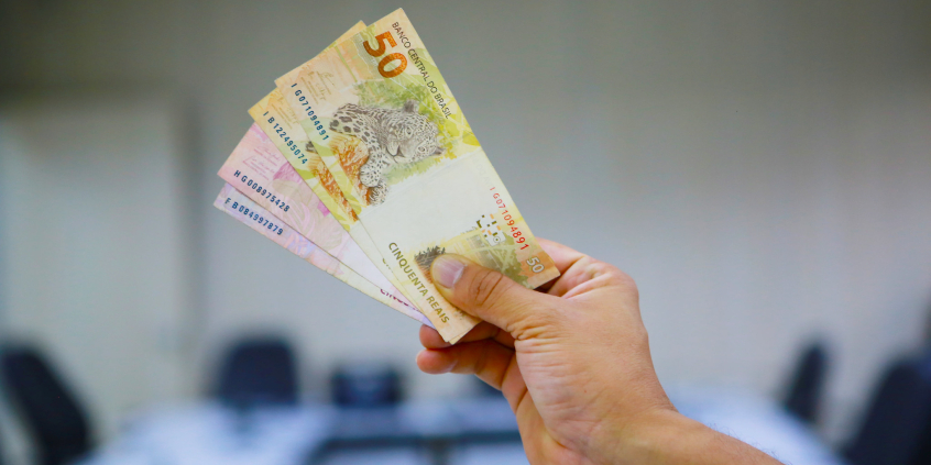 Imagem da mão de uma pessoa caucasiana segurando notas de 50 e de 10 reais