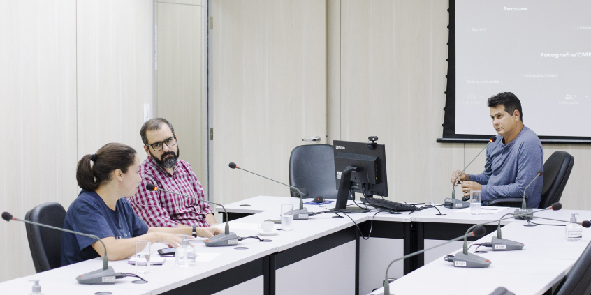 Dois homens e uma mulher, sentados à mesa, em reunião presencial