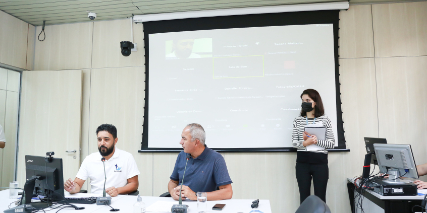 Os vereadores José Ferreira e Claudio do Mundo Novo estão sentado à mesa de reunião. Ao fundo e no alto, o telão mostra a imagem dos outros participantes da reunião. Em pé, uma mulher  usando uma blusa listrada, calça preta e máscara, segura uma pasta de documentos