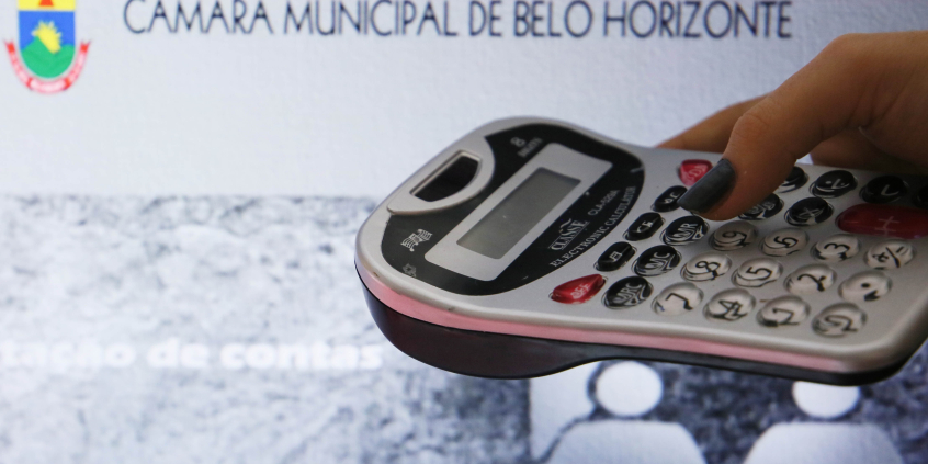 Foto mostra mão segurando calculadora com fundo sobre imagem ilustrativa da CMBHãmara
