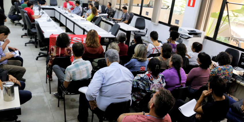 Mais de vinte pessoas sentadas acompanham reunião com oito homens e mulheres sentados à mesa.