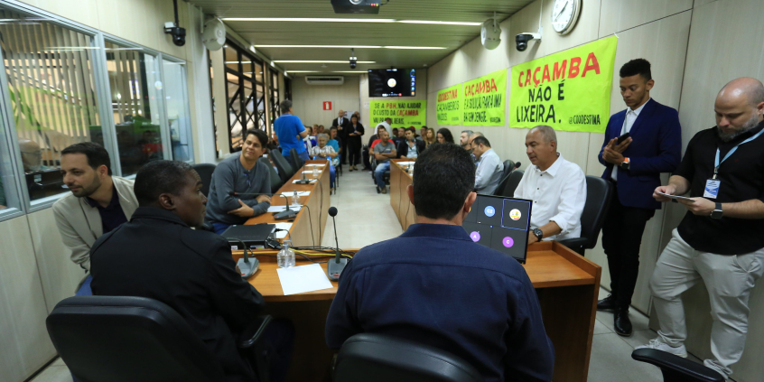 Sala de reuniões com muitos homens e mulheres sentados e de pé, cartazes com dizeres como "Caçamba não é lixo" não paredes.