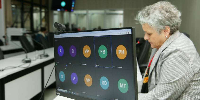 Imagem da tela do computador com indicação de participantes on line. ao fundo, a vereadora Cida Fallabela participa presencialmente 