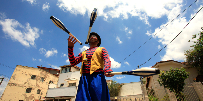 Homem com pernas de pau e vestido de palhaço faz malabares em dia ensolarado.