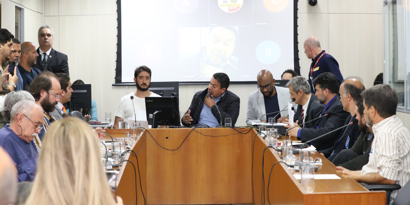 Seis parlamentares e quatro pessoas sentadas à mesa, em uma sala cheia de cidadãos, durante audiência pública.