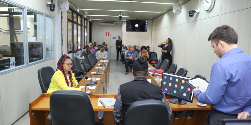 Dois parlamentares reunidos, sentados à mesa, auxiliados por um assessor técnico da câmara, de pé, ao lado deles. ao fundo, mais de dez pessoas assistem à reunião, sentadas.