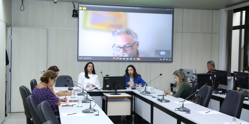 Cinco mulheres reunidas, sentadas à mesa. Em telão ao fundo, representante da Prefeitura participa de reunião de maneira remota.
