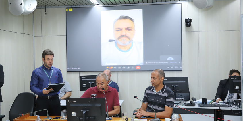 Dois parlamentares sentados à mesa, em reunião. Ao fundo, parlamentar que participa da reunião de maneira remota tem sua imagem exibida em telão.