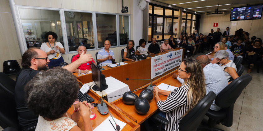 Dose pessoas reunidas, sentadas à mesa, e mais de vinte sentadas ao fundo.