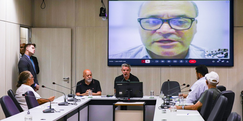Foto de reunião, com convidados sentados à mesa e uma pessoa no telão na parede atrás.