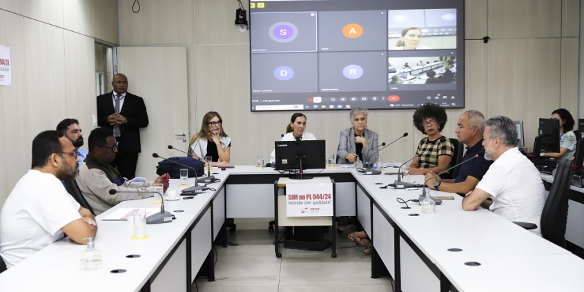 Foto de reunião conjunta. Vereadores estão sentados à mesa com um telão na parte de trás.