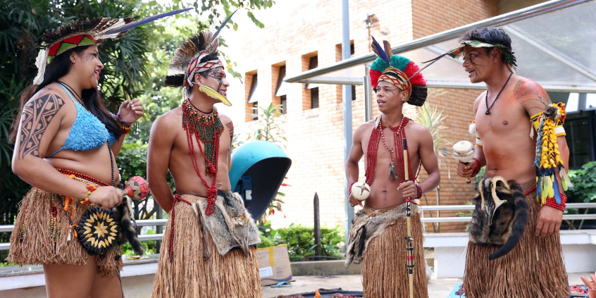 Pessoas indígenas tocam instrumentos de percussão em vestes tradicionais na frente da entrada da CMBH