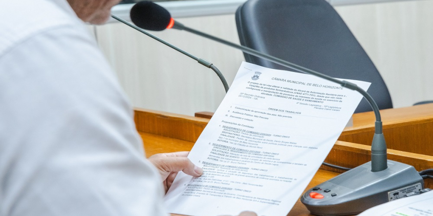 Na foto aparece parte da lateral do braço do vereador, que segura um  papel em que está a pauta da reunião e fala ao microfone.