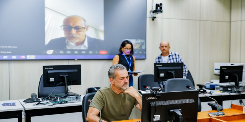 Vereador Wilsinho da Tabu sentado em frente ao computado, com a tela atrás projetando a imagem do vereador Fernando Luiz.