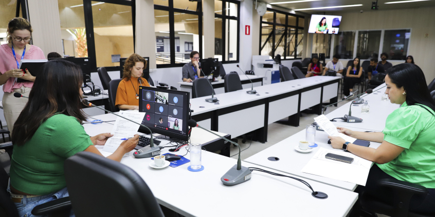Vereadoras assentadas no Plenário Helvécio Arantes, em mesa com formato de U. O computador à frente de Loíde mostra chamada virtual e pequena plateia assiste à frente.