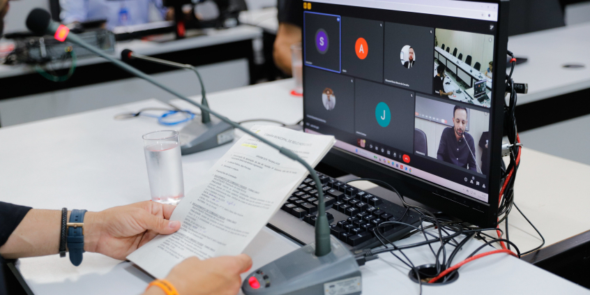 Tela do computador mostra reunião virtual dos vereadores. Em frente à tela, duas mãos seguram um documento com a pauta da reunião.