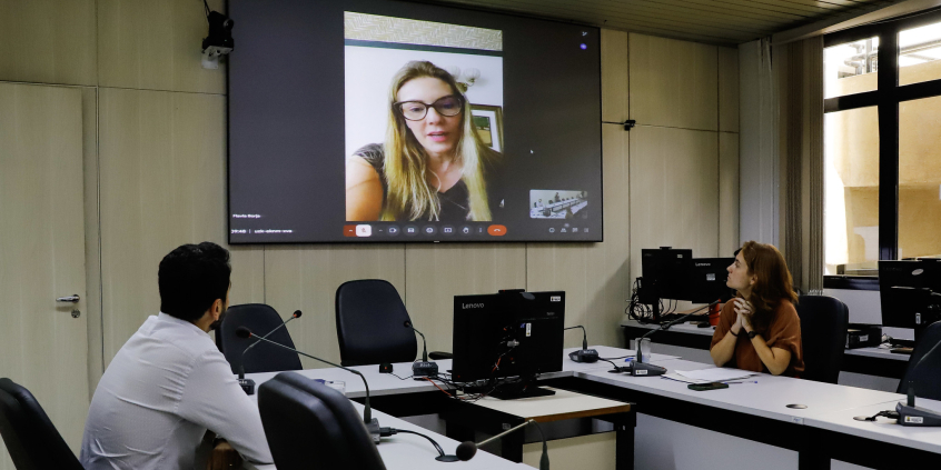 Luiza Dulci e Uner Augusto estão assentados no Plenário Helvécio Arantes. O telão à frente deles mostra Flávia Borja em chamada virtual.