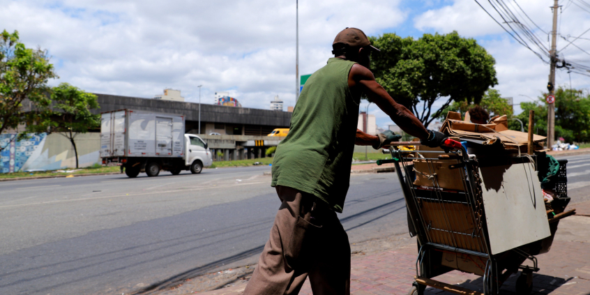 Morador de rua de costas, empurrando carrinho com seus pertences na Praça do Peixe