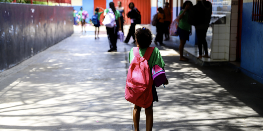 Menina é vista de costas dentro de escola usando uniforme e mochila rosa