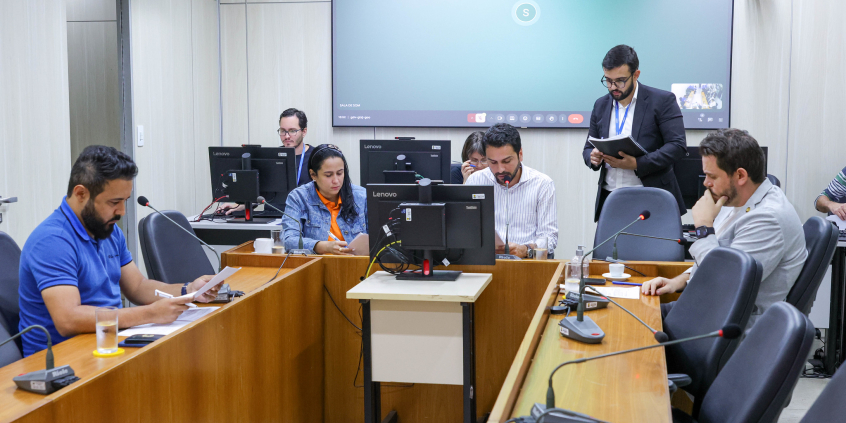 Imagem dos vereadores em reunião sentados em volta de uma mesa em formato U com um telão azul ao fundo