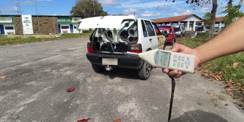 carro com som automotivo instalado no porta malas; close nas mãos de pessoa com instrumento de aferição de ruído