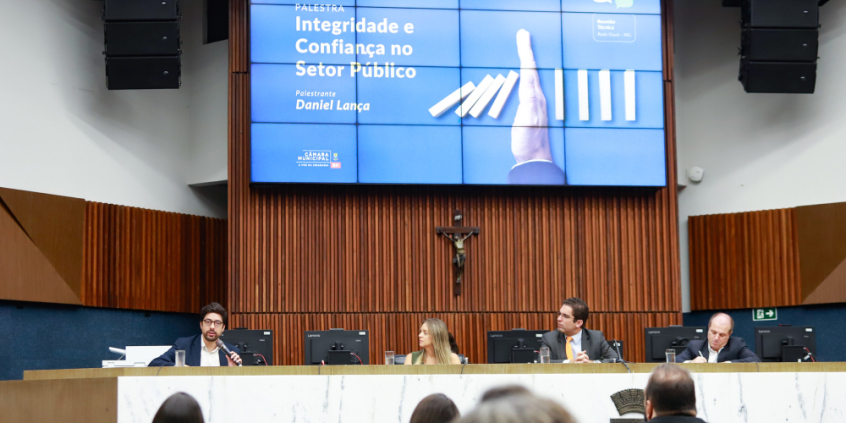Mesa do Plenário Amintas de Barros coma  presença do vereador Braulio Lara e outros participantes. No telão, está o escrito "Palestra Integridade e Confiança no Setor Público, palestrante Daniel Lança".