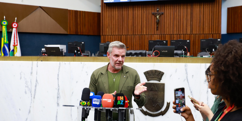 Foto do vereador Professor Juliano Lopes, que aparece centralizado falando com repórteres  