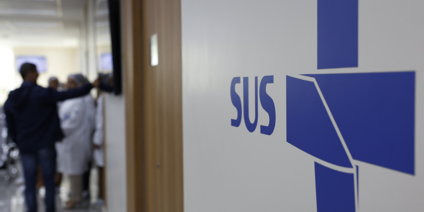 Imagem de corredor de unidade de saúde, com a inscrição SUS na parede