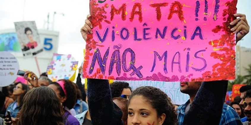 Mulher segura cartaz com os dizeres: "Seu machismo mata!!! Violência não mais"