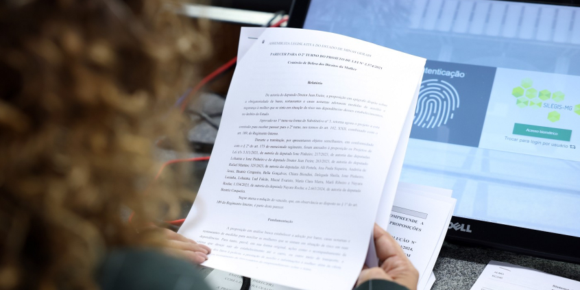 mulher de costas segura folha com texto em frente a tela de computador com imagem sobre identificação digital