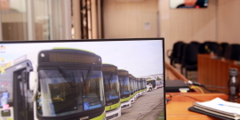 monitor de computador exibe foto de ônibus estacionados. Ao fundo, plenário vazio e telão com vereadores e convidados em videoconferência