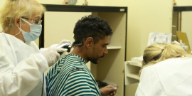 Duas profissionais de saúde com máscara oferecem cuidados a um homem, uma delas corta o cabelo dele. Visita técnica da Comissão de Saúde ao CERSAM - Leste
