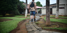 Homem jovem, de costas, percorre um caminho em local com casas, árvores, grama e terra, durante o dia. 