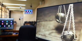 Imagem  dois pêndulos na cor prata refletida na tela do computador, fazendo alusão à balança da justiça. Ao fundo a imagem desfocada da sala de reunião