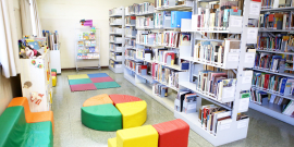 Biblioteca infantil, com bancos e tapetes coloridos. Ao centro, mesas formada por quatro triângulos coloridos